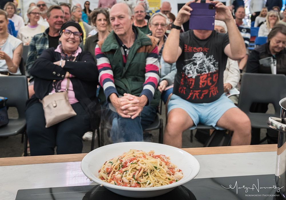 A plate of pasta in front of a crowd. Person covers face with purple item. Others watch and smile.