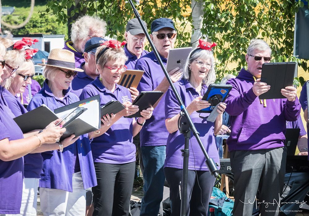 Choir in purple shirts performing outdoors, holding songbooks and tablets.