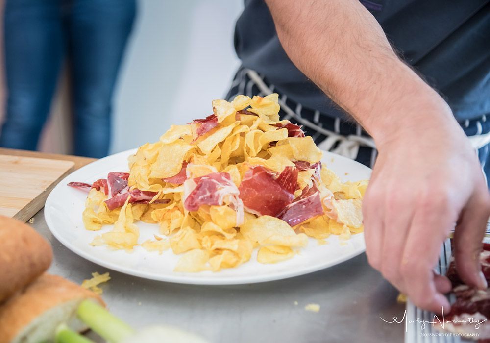 Person arranging potato chips and ham on a white plate in a kitchen.