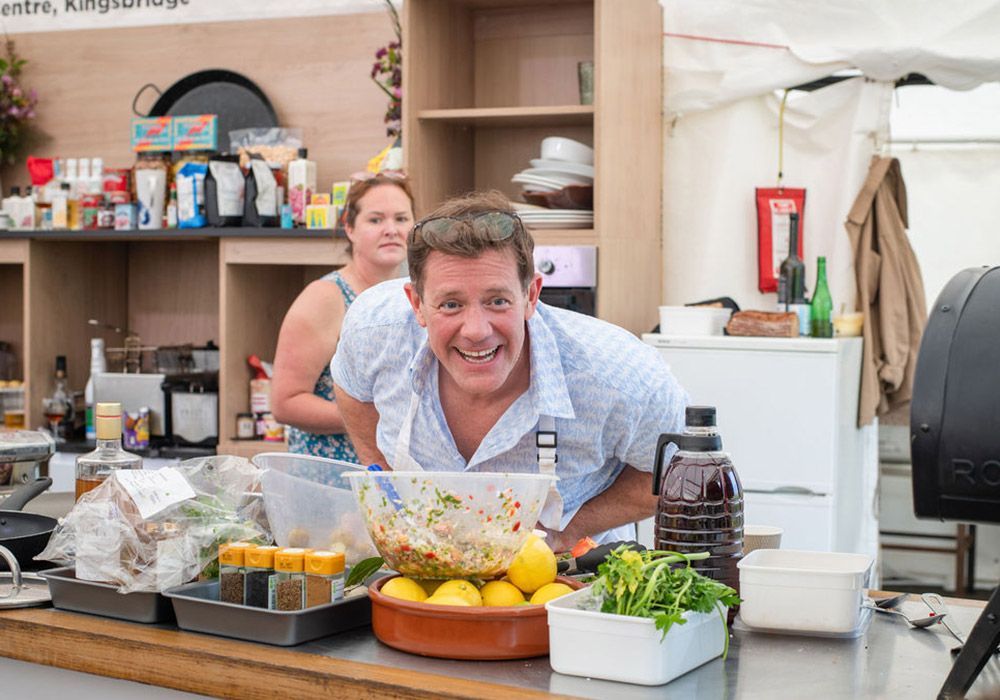 Chef smiles while prepping food at outdoor cooking event, assistant behind him.