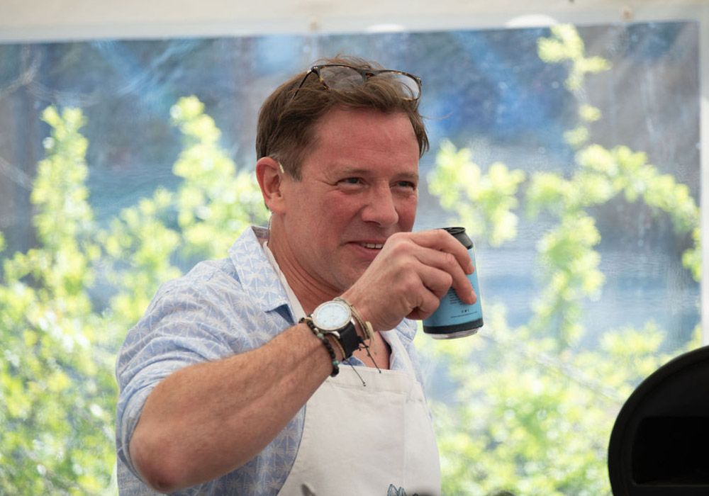 Man in apron smiles, holding a blue can, outdoors near greenery.