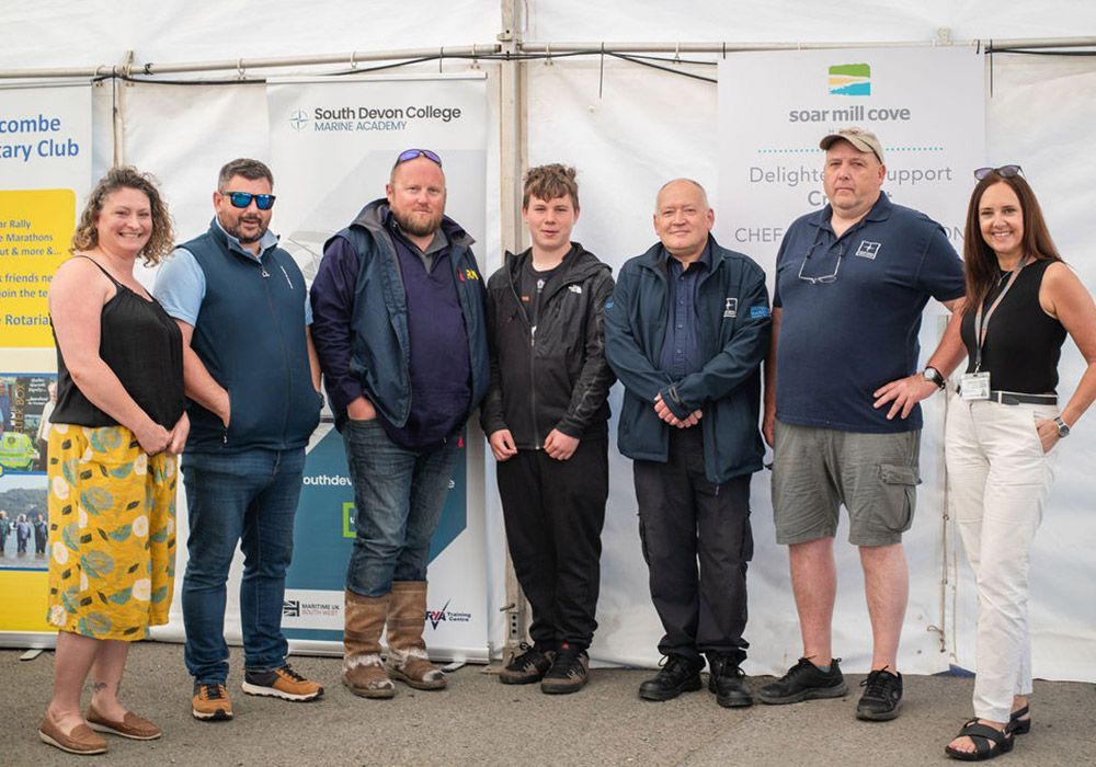 Group of seven people standing in front of a white tent, smiling. The background has business signage.