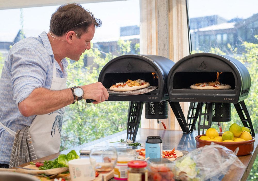 Man tending to pizzas cooking in two black outdoor pizza ovens, sunny day.