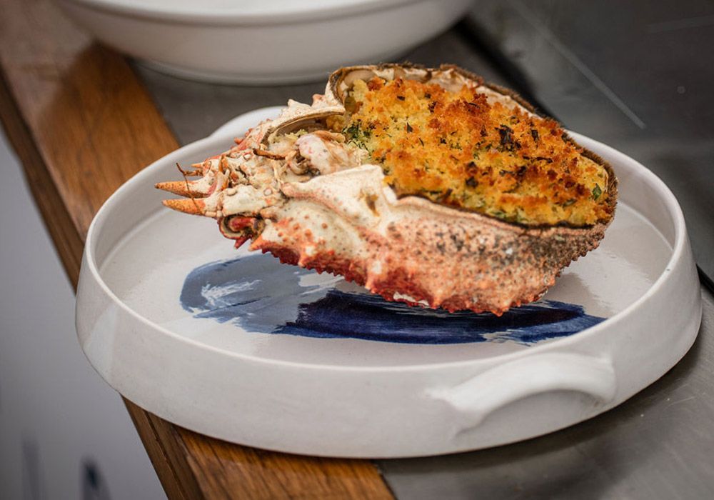 Crab dish on white plate with blue brushstroke. Crab shell filled with stuffing, resting on a wooden surface.