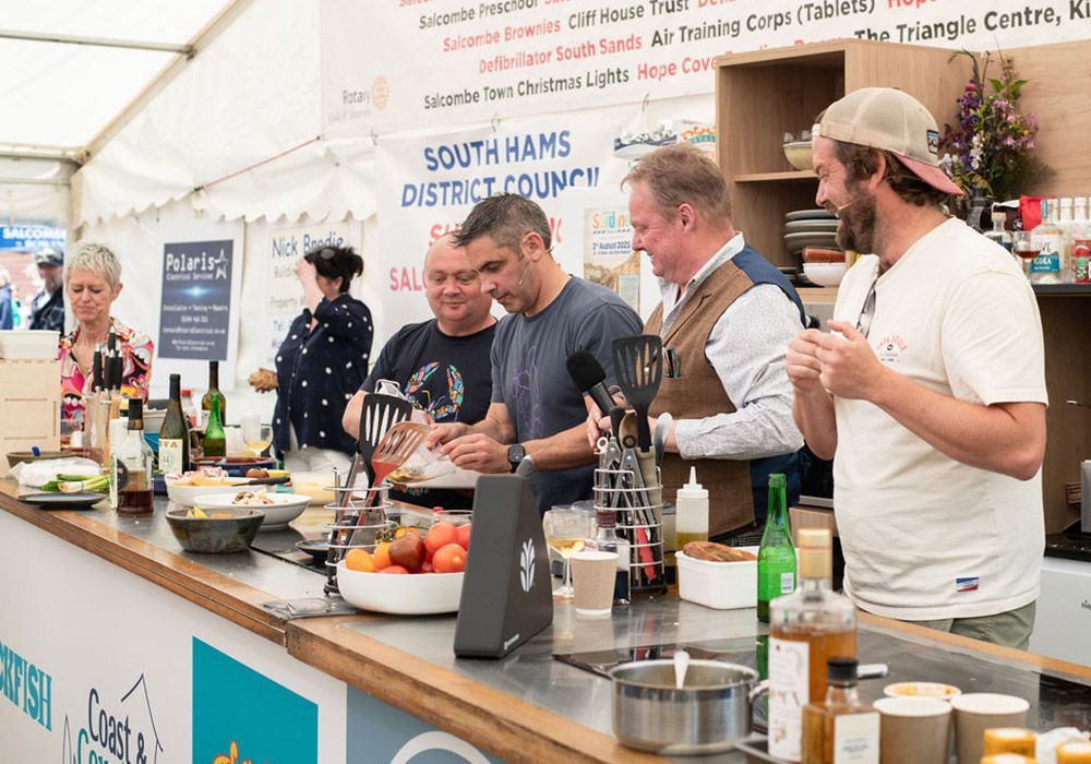 Chefs cooking at a food festival:  men laughing behind a counter, fresh ingredients, white tent in the background.