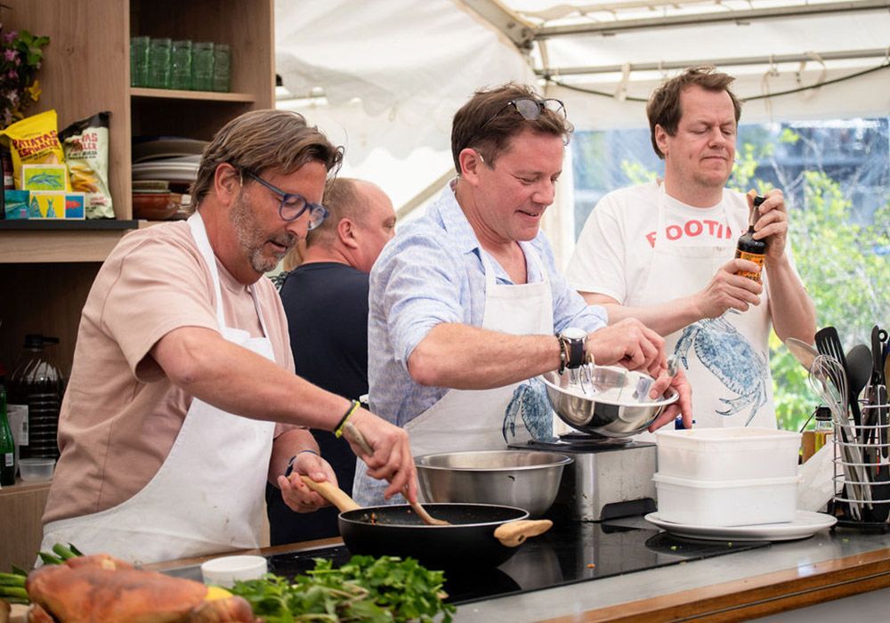 Three men cook together in a kitchen tent. One stirs a pan, another mixes a bowl, and the third pours from a bottle.