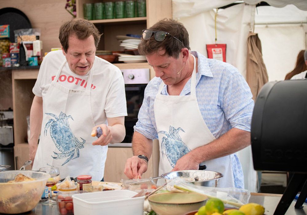 Two men cooking in a kitchen, both wearing aprons with blue crab design.