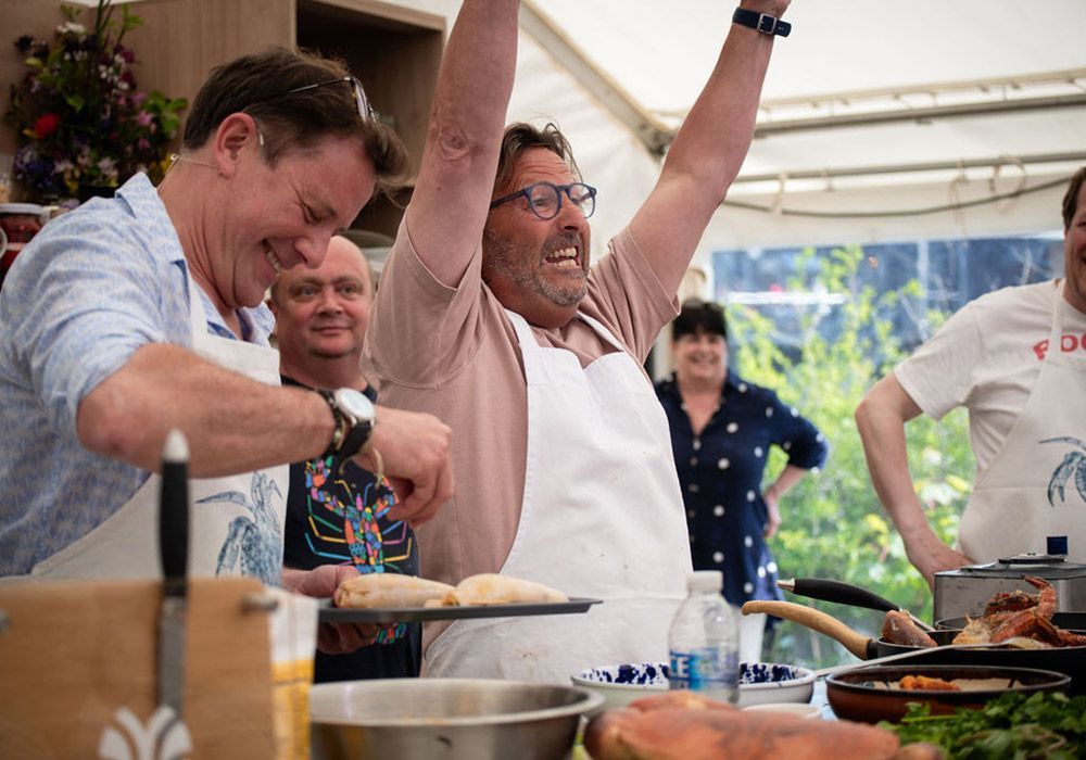 Man celebrates cooking success; others smile and watch. Tent setting, food prep visible.