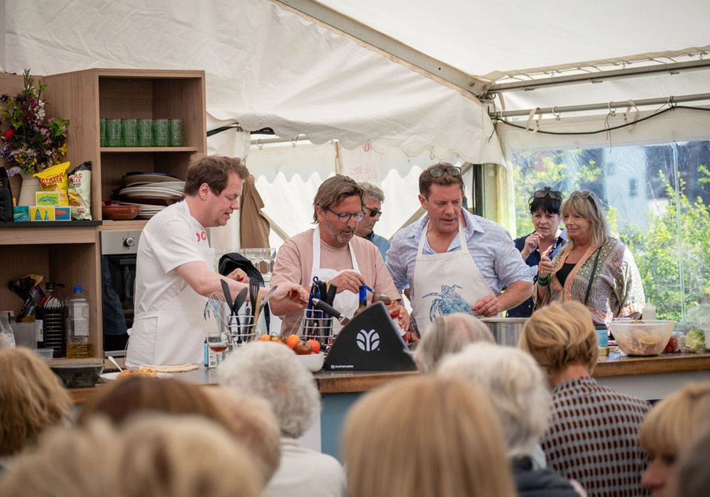 Chefs demonstrate cooking at an outdoor event. Audience watches, white tent overhead.