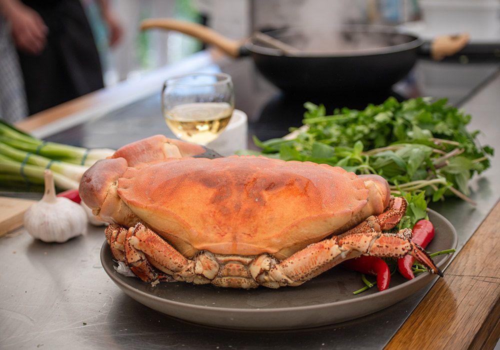 Cooked crab on a plate with herbs, garlic, and chilies. A glass of wine and a pan are in the background.