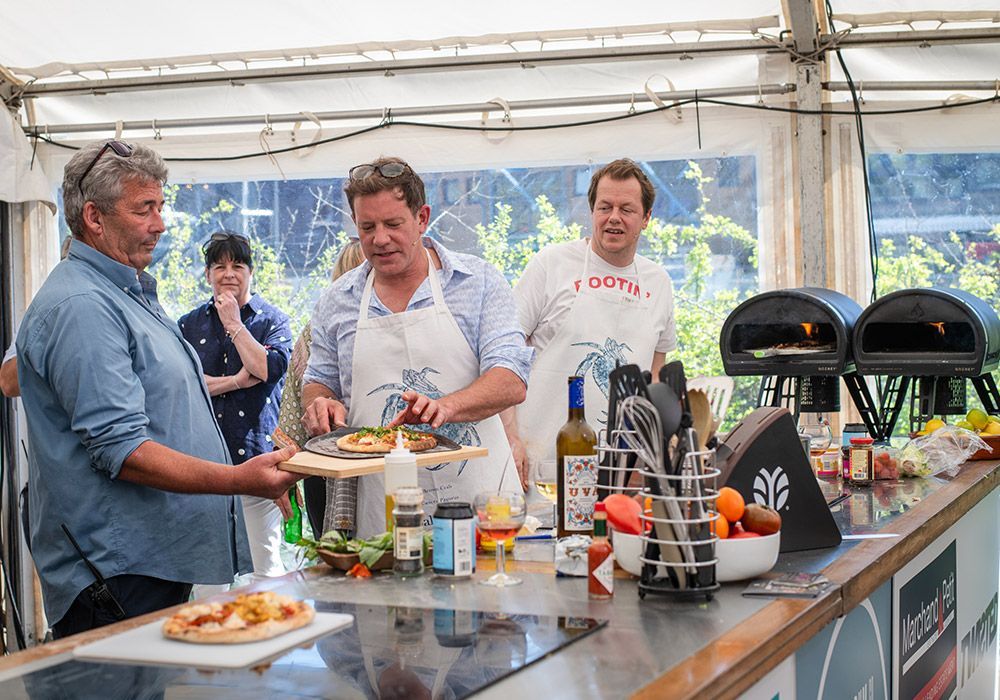 Chefs preparing food at an outdoor cooking demonstration, with pizza ovens and ingredients visible.