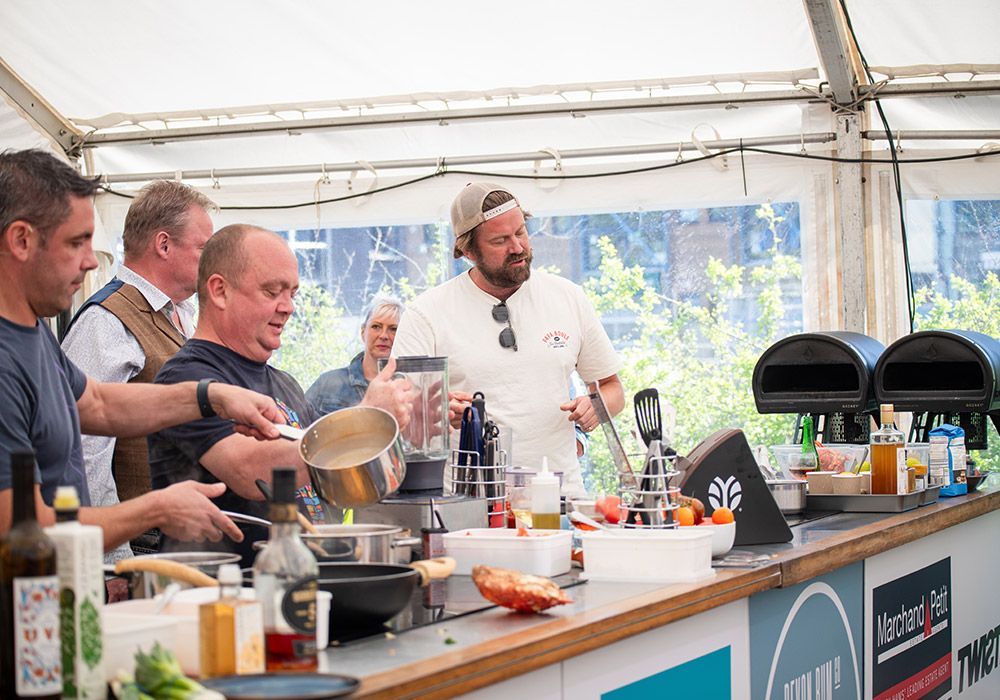 Chefs cooking in a tent, stirring pots and pouring ingredients, sunny day.