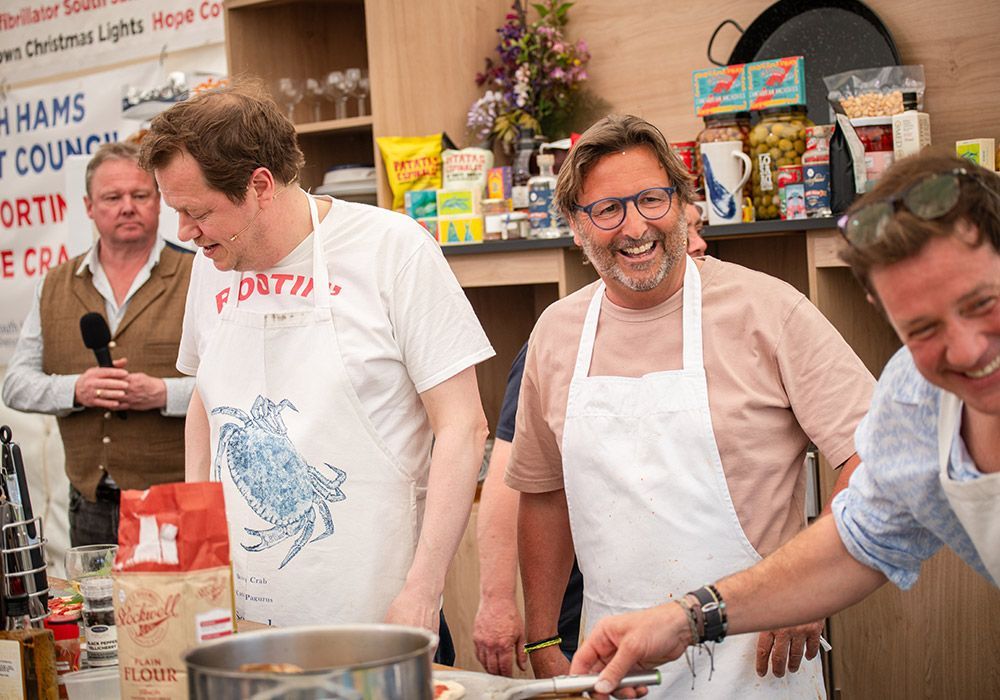 Three men wearing aprons, smiling, and cooking at an outdoor event. A fourth man holds a microphone nearby.