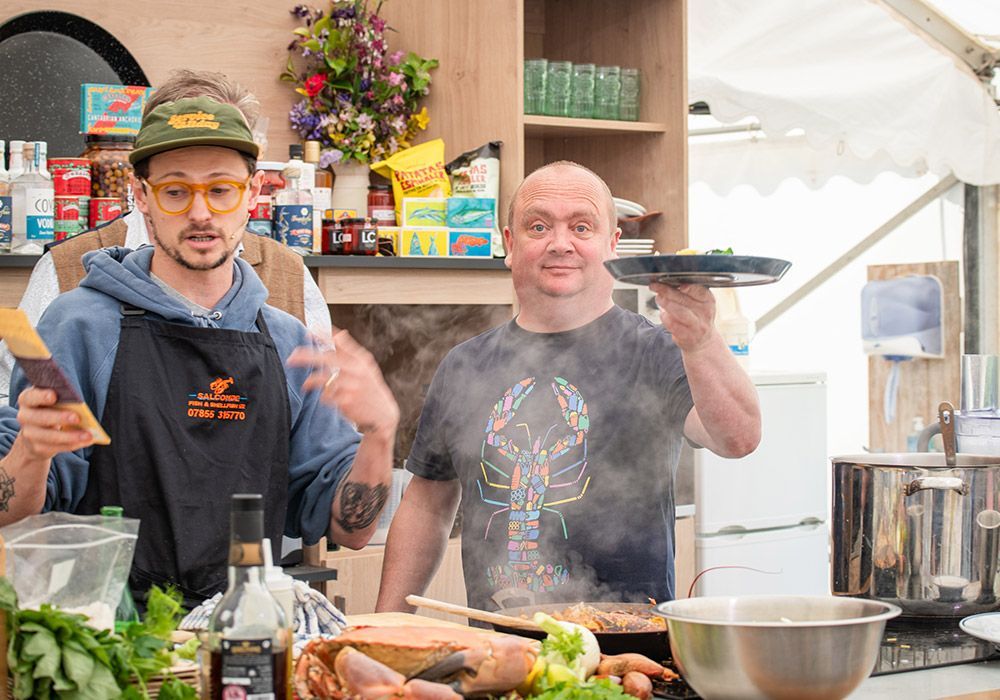 Two men cooking, one holding a plate, the other gesturing. Kitchen with ingredients and steam.