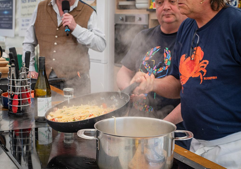 Three people cooking, one with microphone, pasta in pan with steam, pot below.
