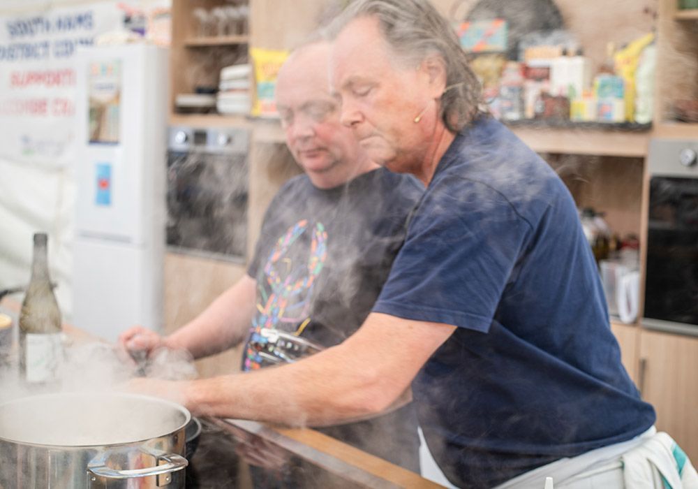 Two men cooking, leaning over a steaming pot, in a small kitchen.