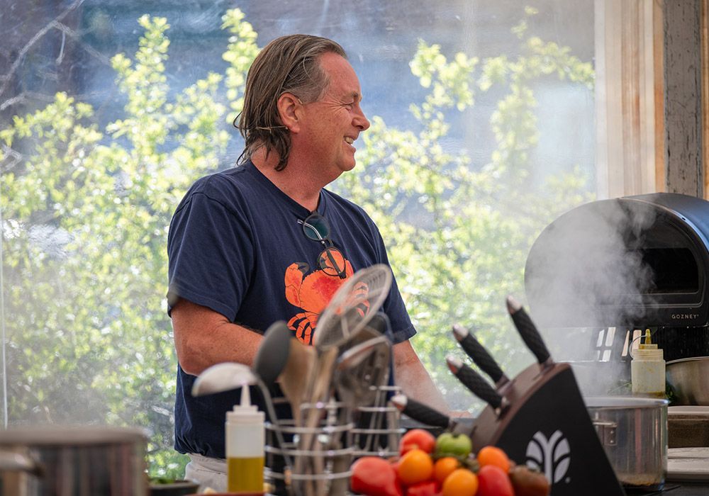 Man in a blue crab shirt smiles while cooking with steam, surrounded by cooking tools and food.
