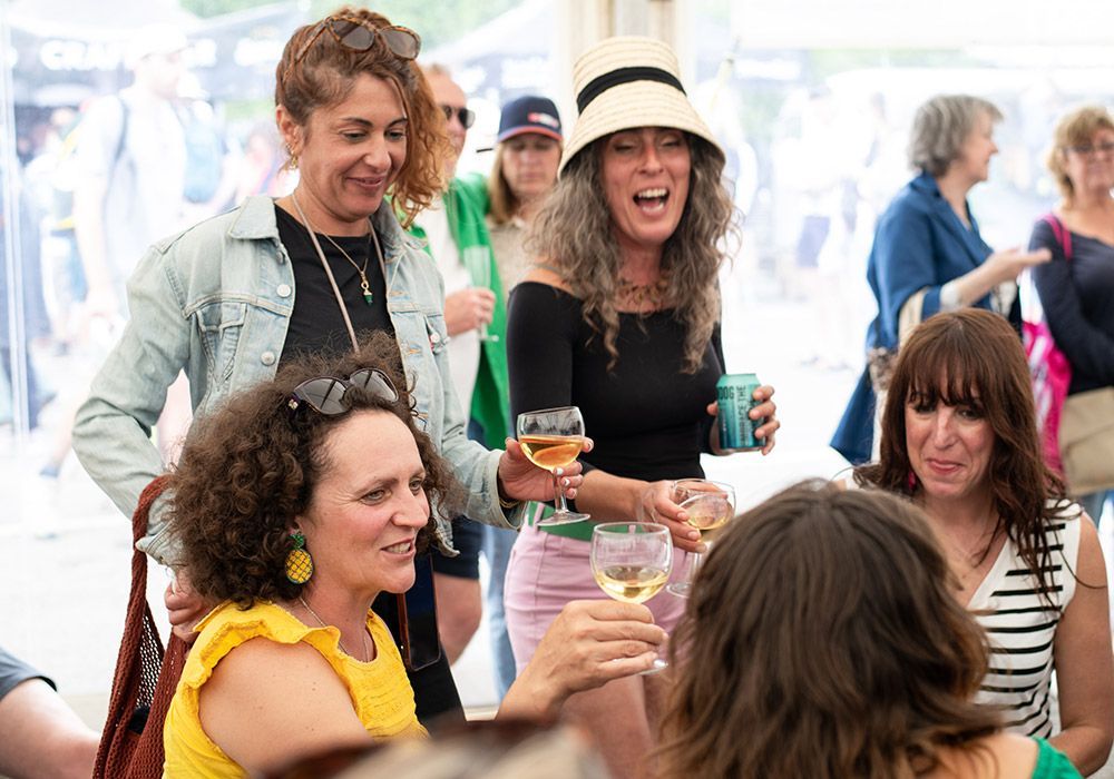 Group of women at an outdoor event, drinking wine and smiling. One wears a sun hat and denim jacket.