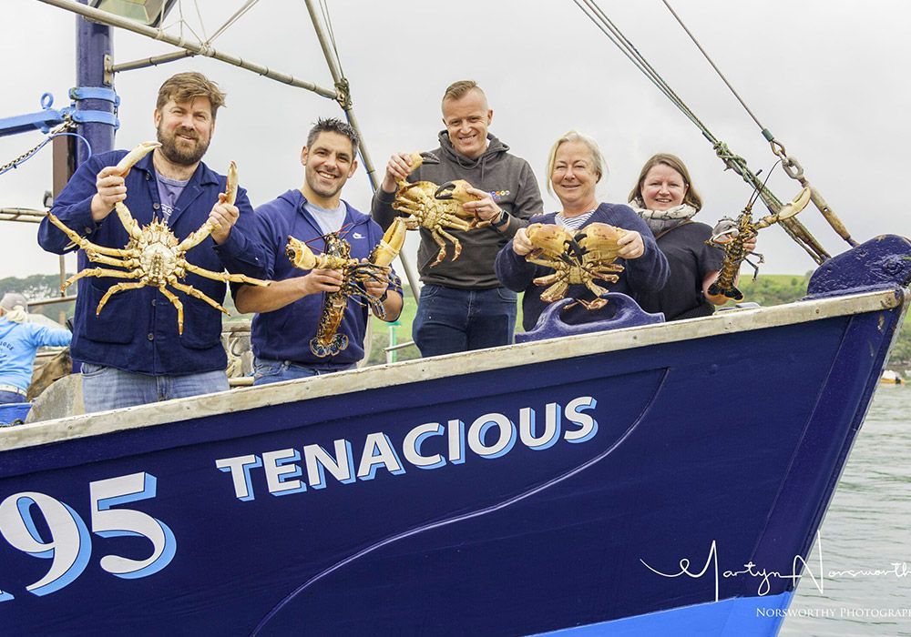Five people on a boat, holding up crabs and lobster. The boat is blue, named