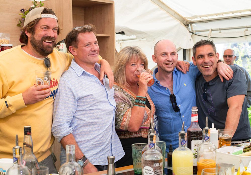 Five people smiling, arms around each other, at an outdoor bar with bottles and drinks.