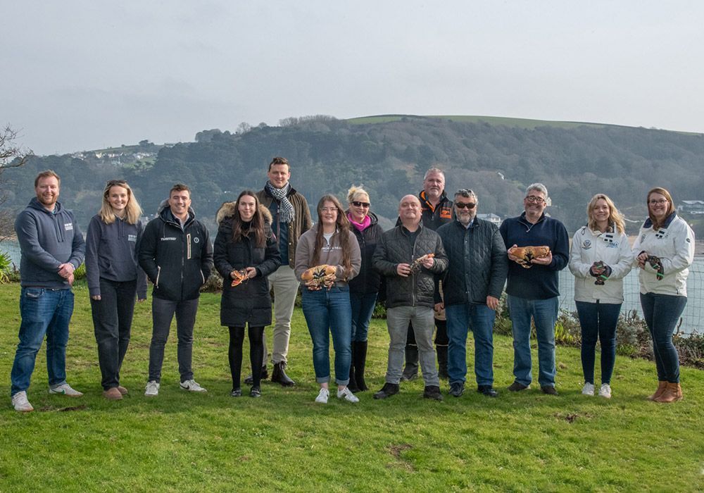 Group of people outdoors, holding small dogs, standing on grass by water with trees in the background.