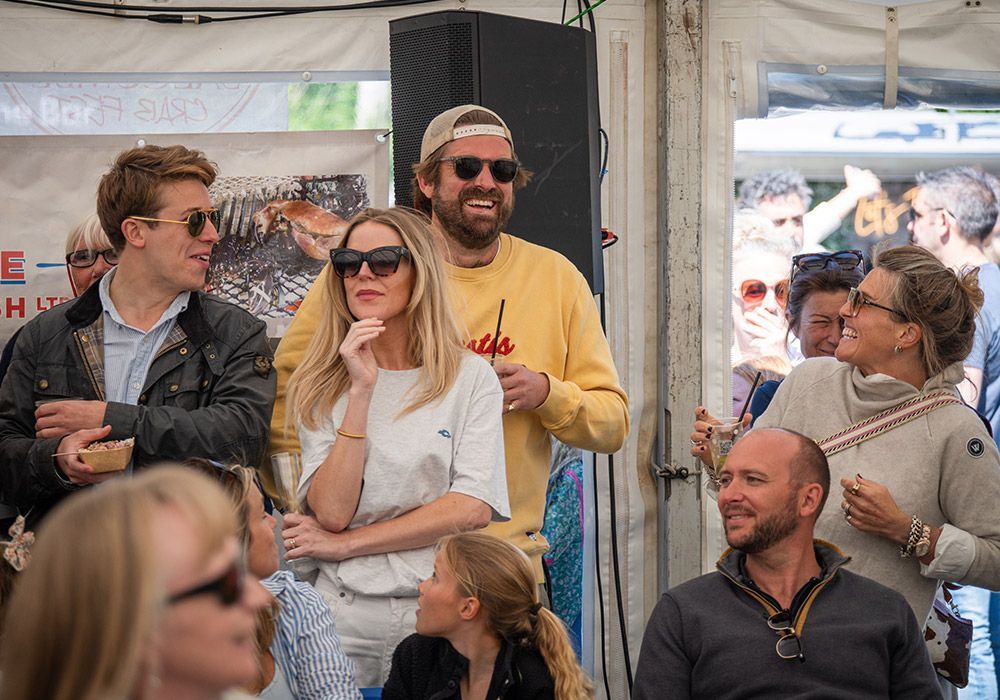 People gathered outdoors, some smiling, wearing sunglasses. Man in yellow sweatshirt, woman in white top, listening.