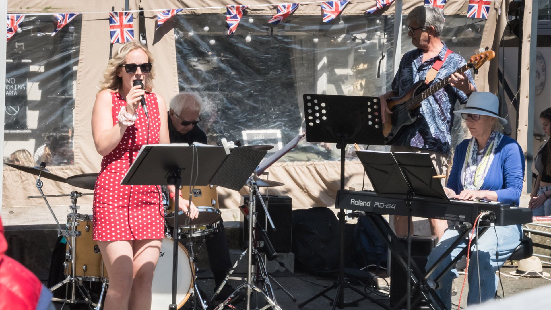 Band playing outdoors under Union Jack bunting. Female singer in red dress, keyboardist, guitarist, and drummer.