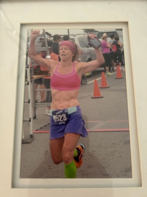 Woman crossing finish line, arms raised, wearing running clothes, pink top, blue shorts, running event.