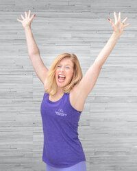 A woman in a purple tank top is standing with her arms in the air.