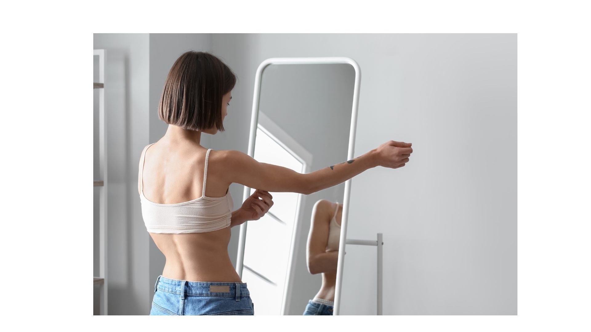 Woman in a crop top and jeans looks at her reflection in a full-length mirror, reaching for her arm.