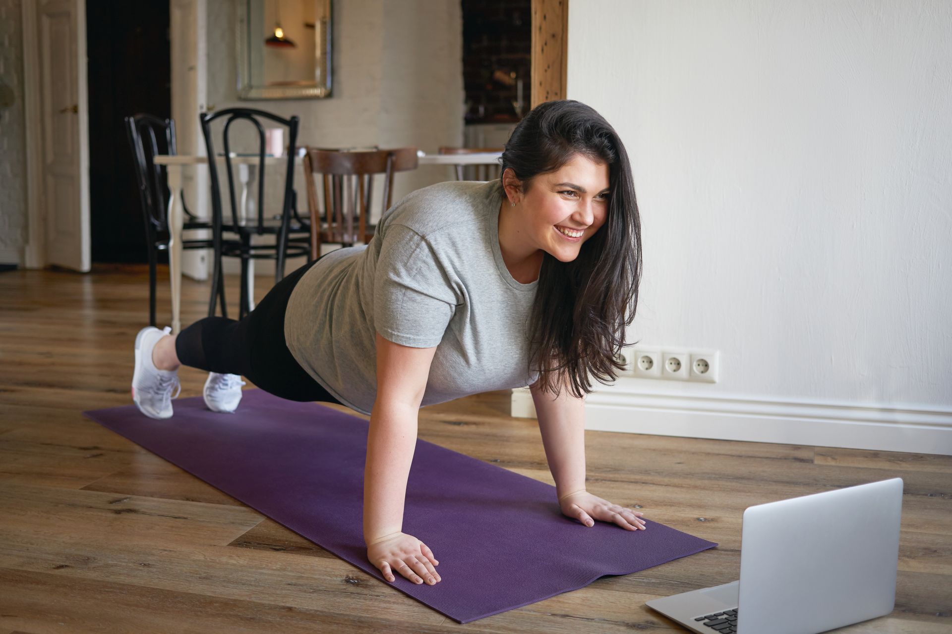 A woman is doing push ups on a yoga mat in front of a laptop.
