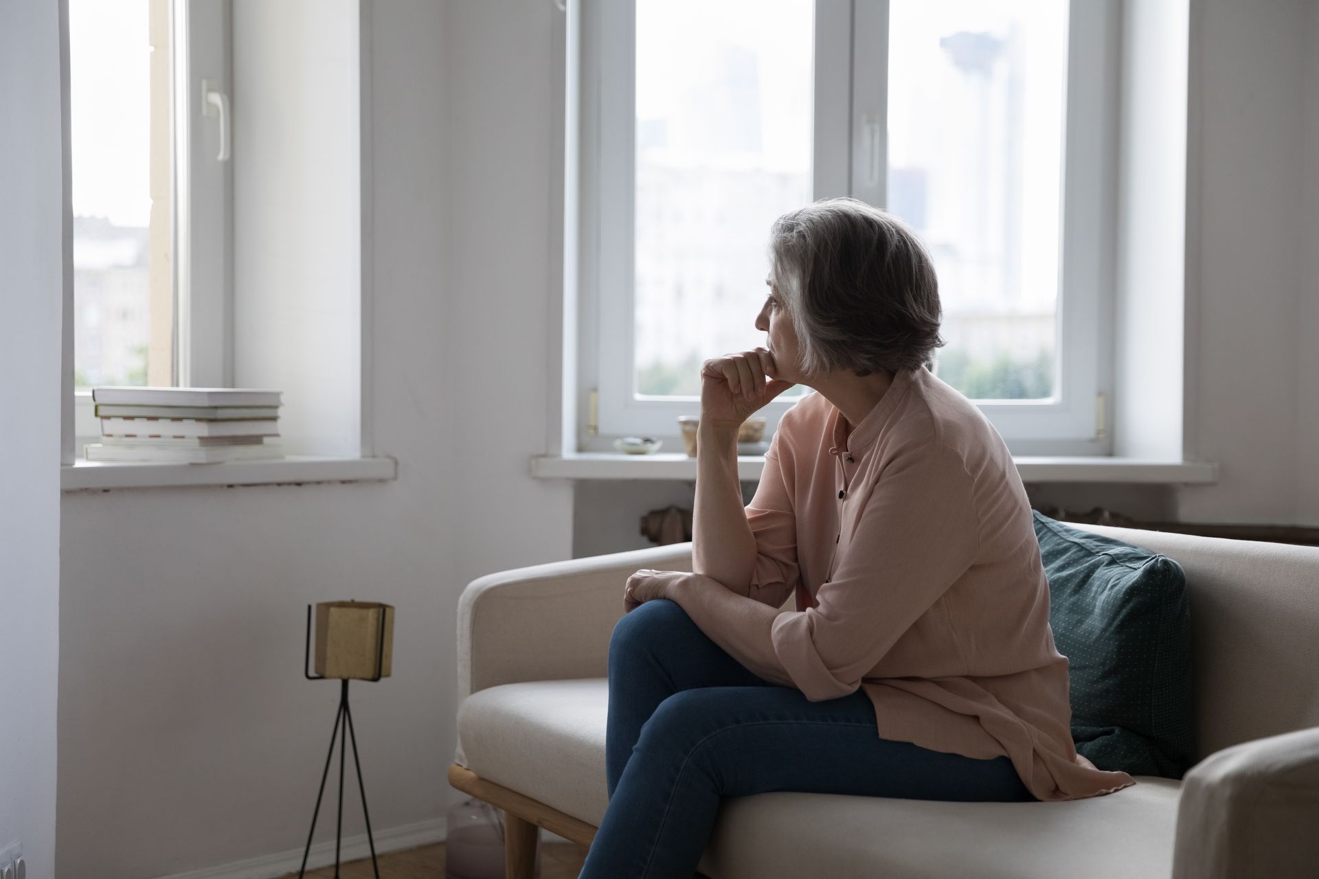 An elderly woman is sitting on a couch looking out a window.
