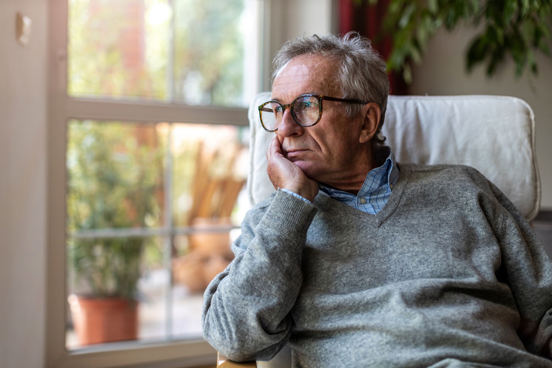 Man in glasses, resting his chin on hand, looking out a window. Sitting in a chair.