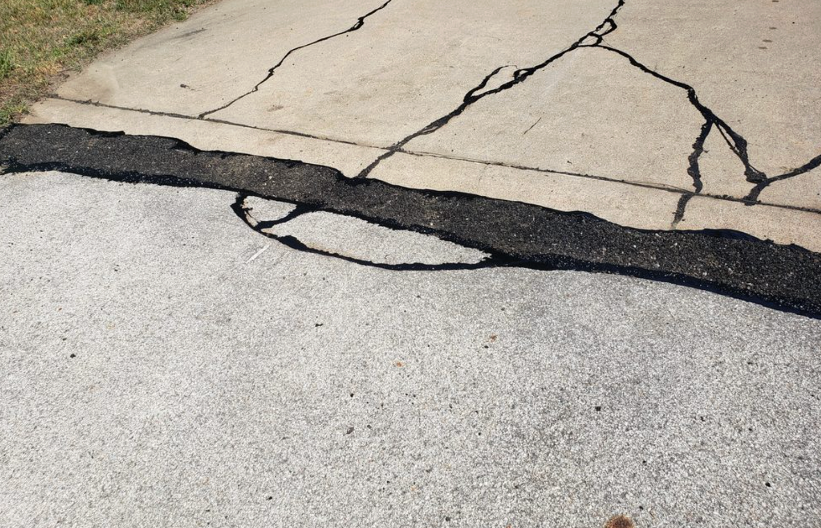 A concrete driveway with a large asphalt patch repair and several deep cracks.