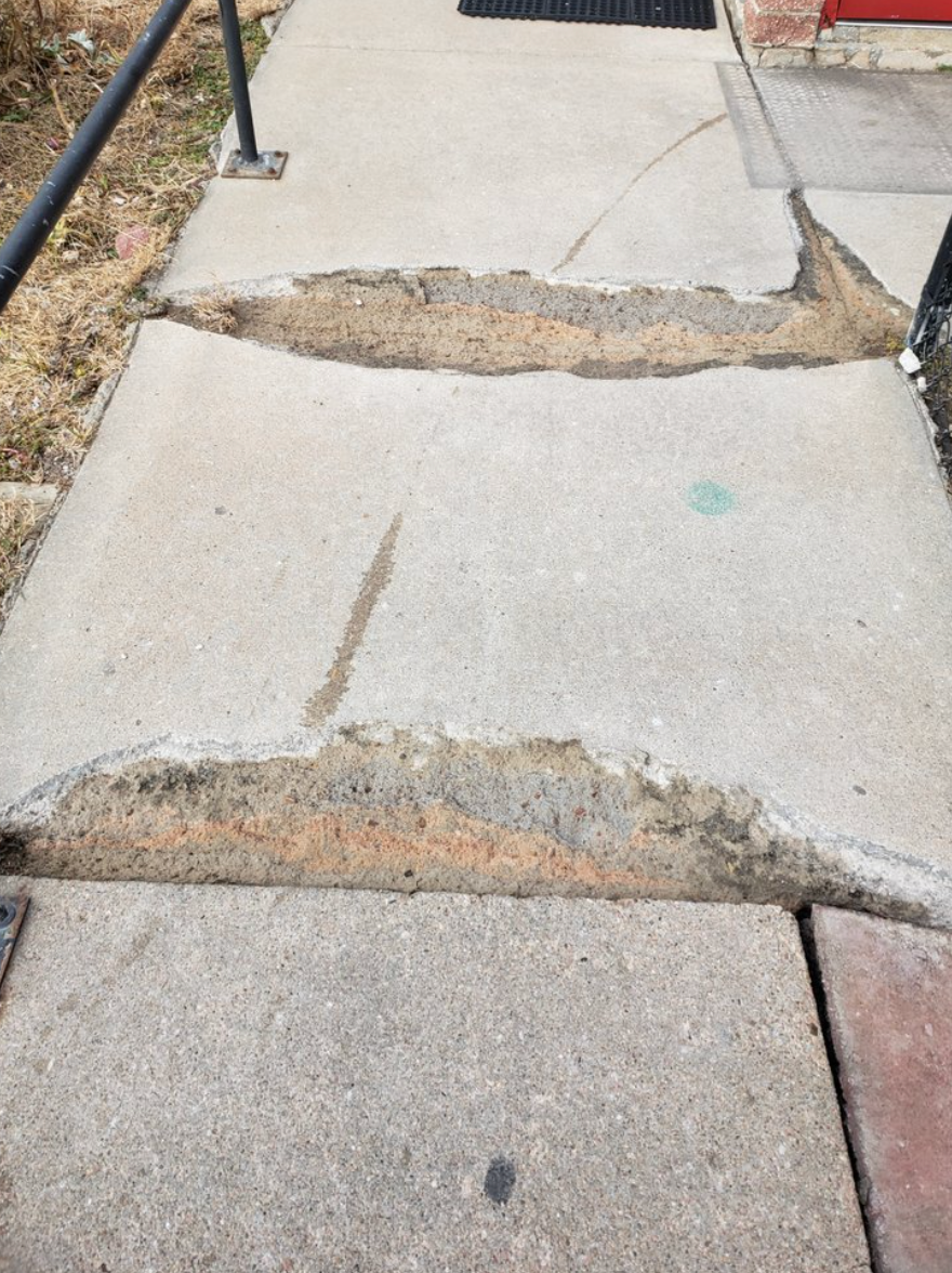 A concrete sidewalk with significant cracks and wide gaps between slabs, posing a trip hazard for pedestrians.