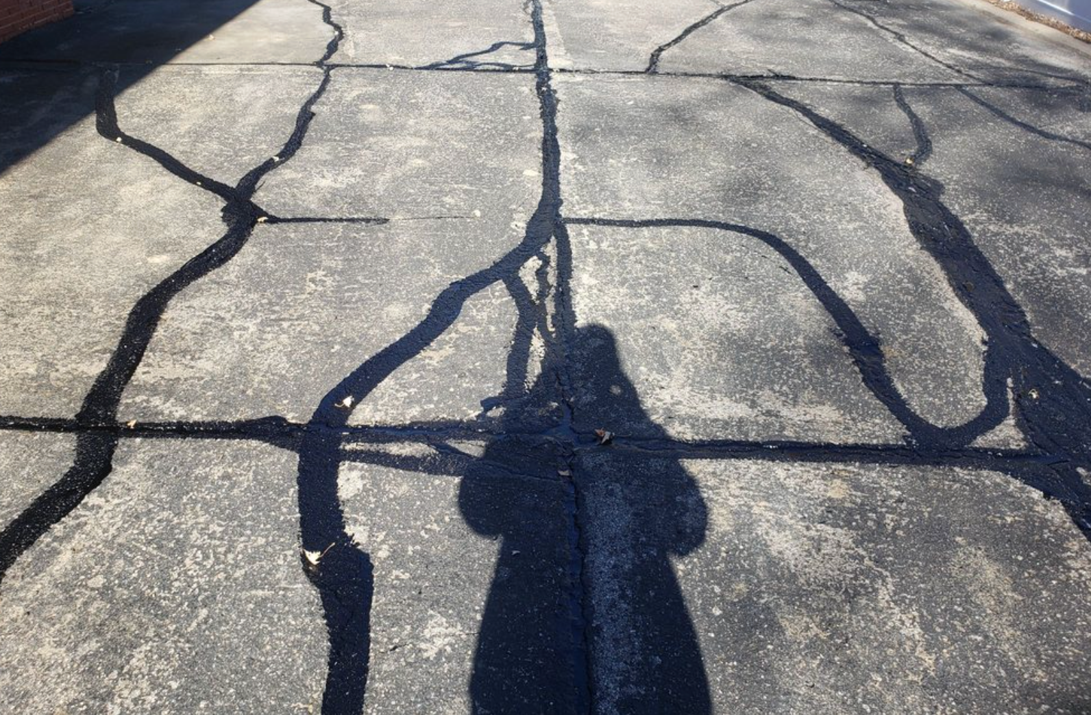 A person’s shadow cast across a paved surface covered in deep, interconnected cracks.
