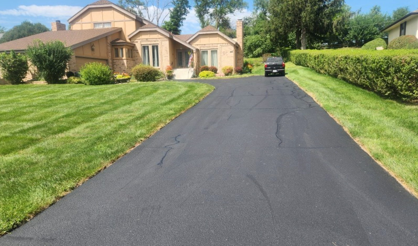 A freshly paved asphalt driveway leading to a suburban house with a green lawn and trimmed hedges on a sunny day.