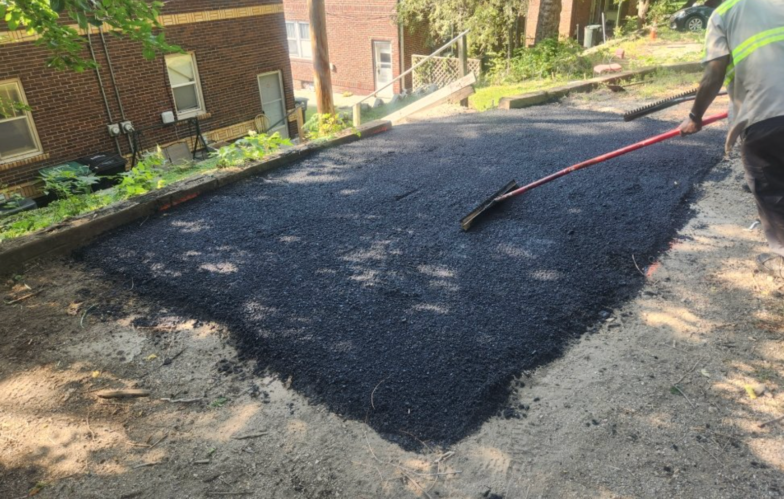 Road workers laying asphalt, one raking, and a roller in background.