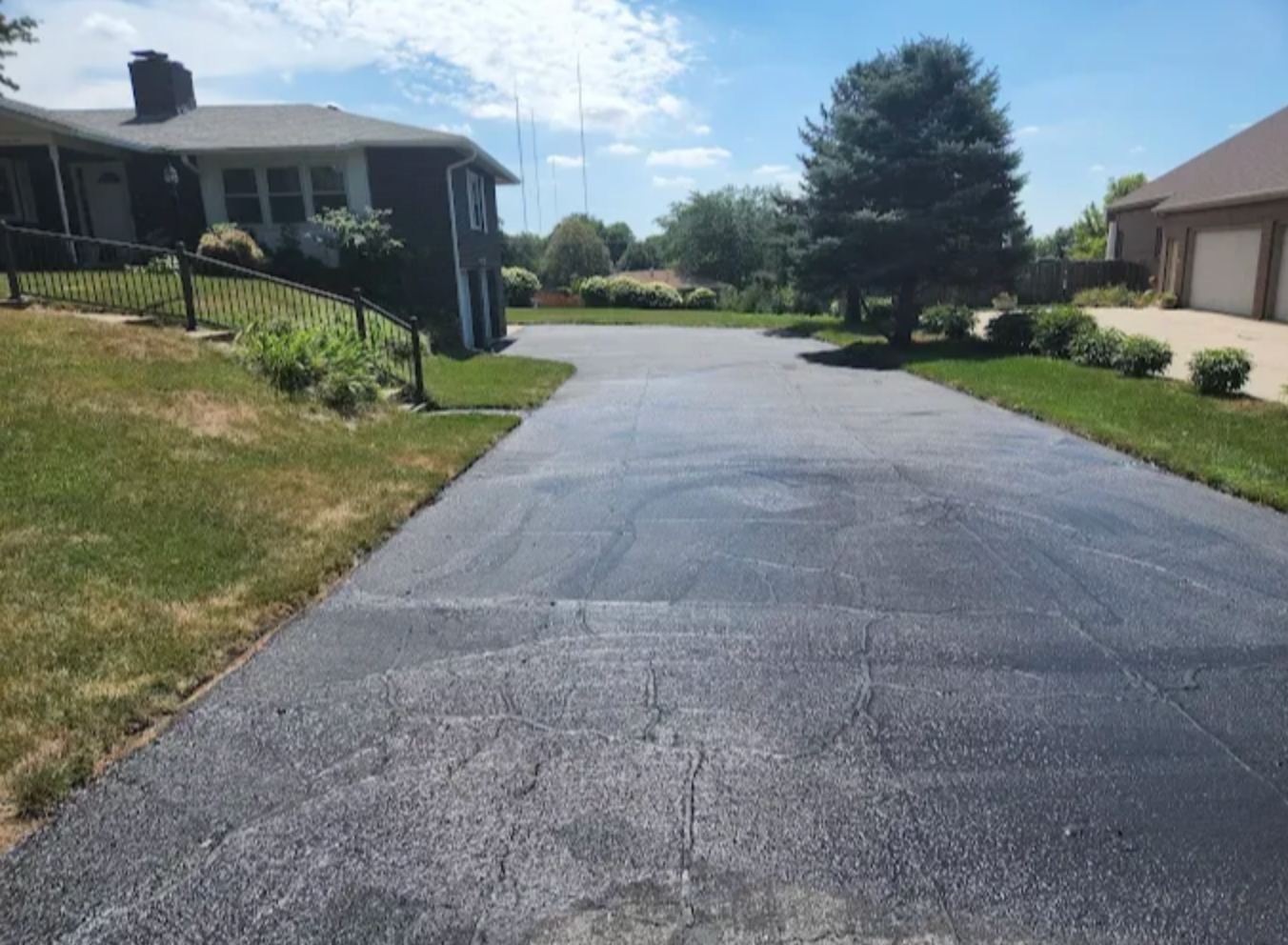 An asphalt driveway leads toward a house and detached garage on a sunny day with green grass and trees.