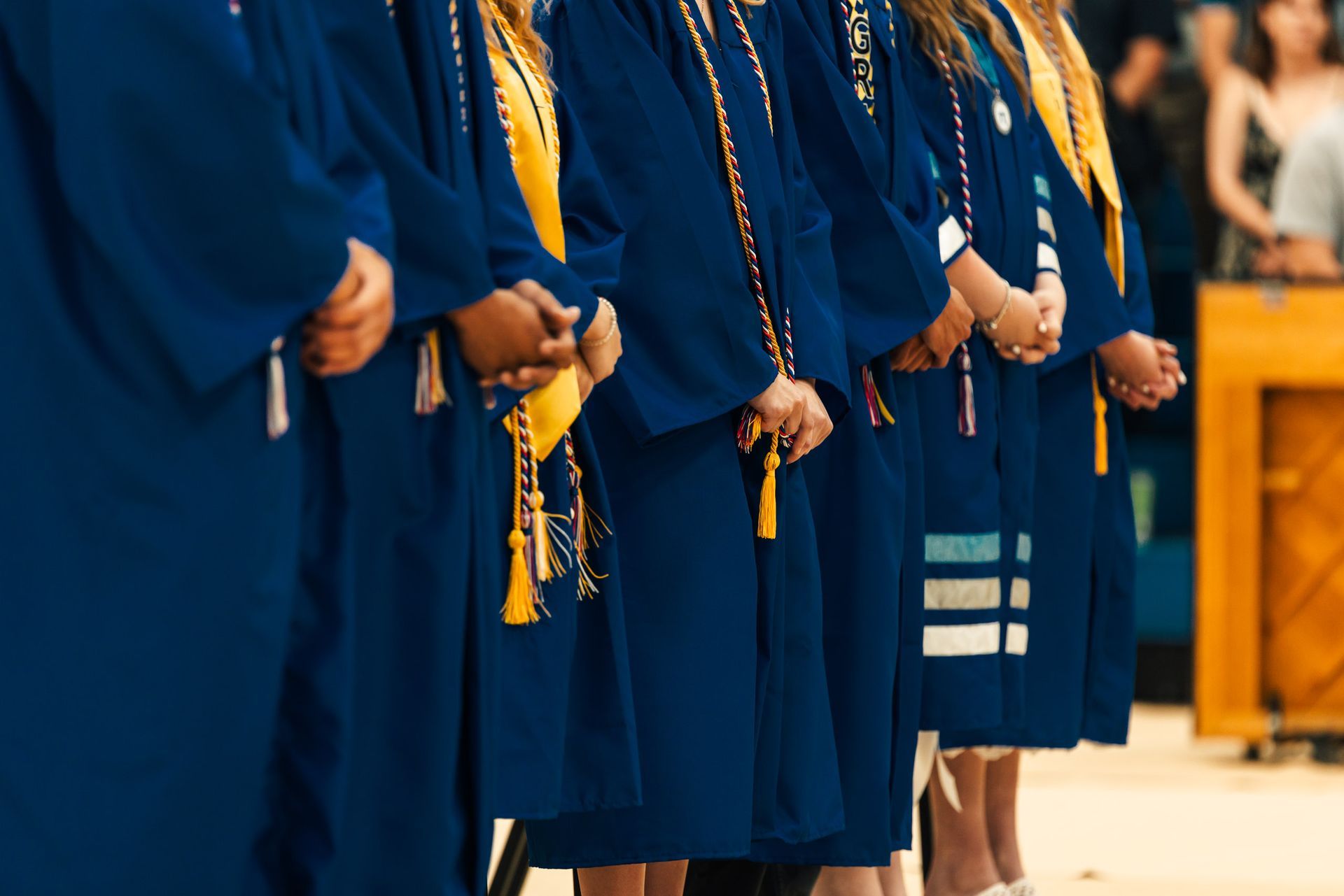 Graduates in blue gowns stand in a row. Some wear stoles, hands clasped.