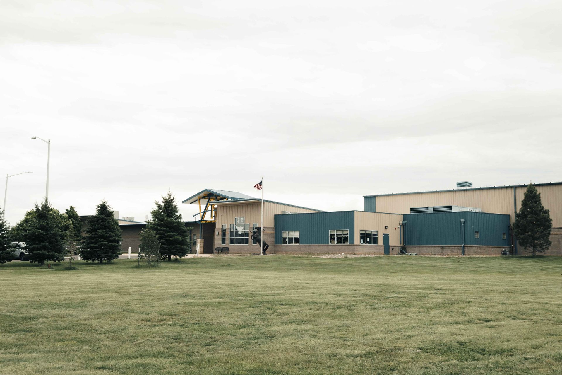 School building with teal and beige walls under cloudy sky. Green lawn and trees in foreground.