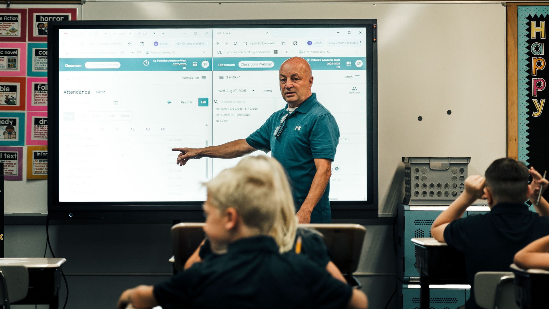 Teacher in blue top speaks in a classroom. Two students stand nearby. American flag hangs behind.