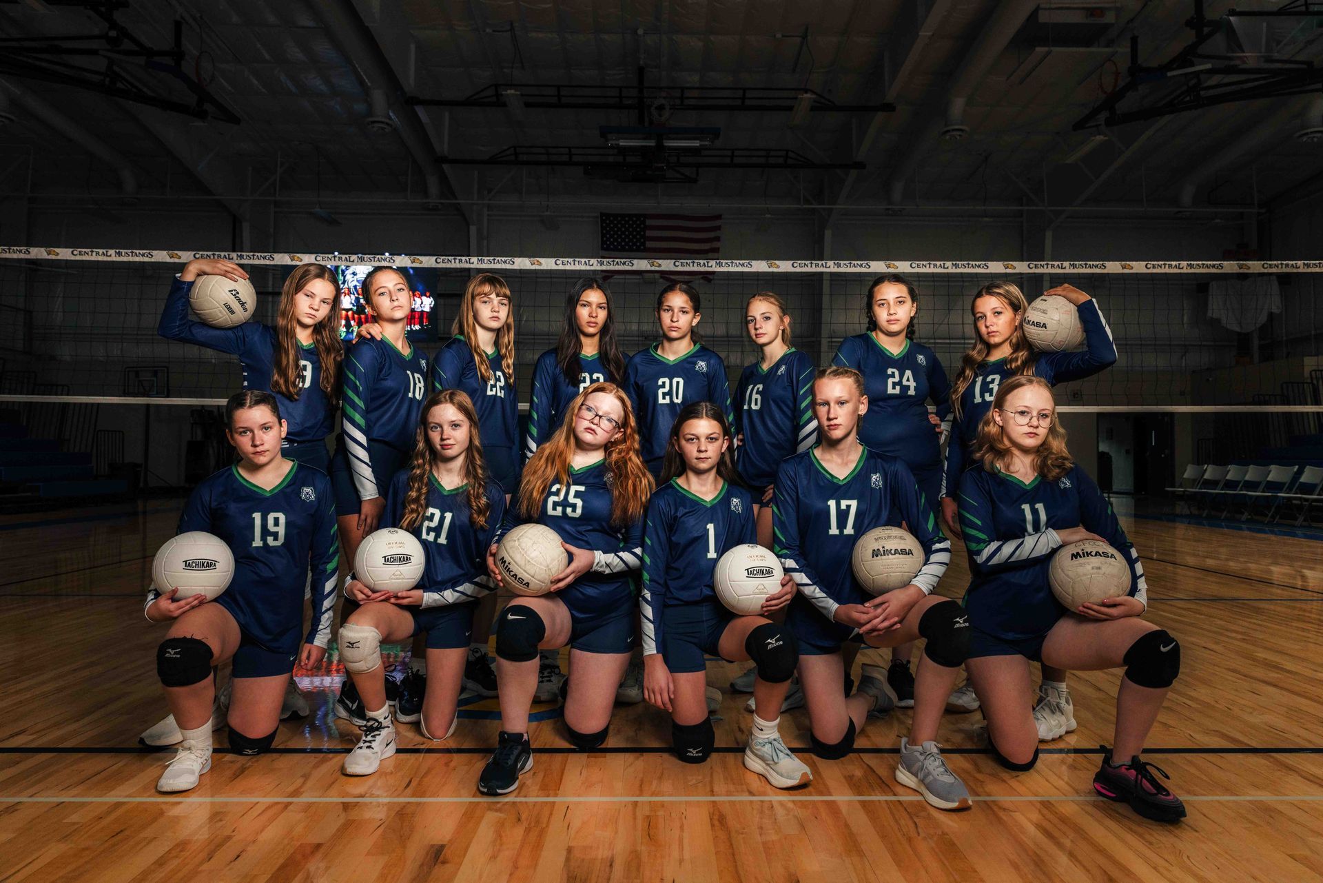 Volleyball team posing for photo, holding balls, in matching blue and green jerseys, in a gym.