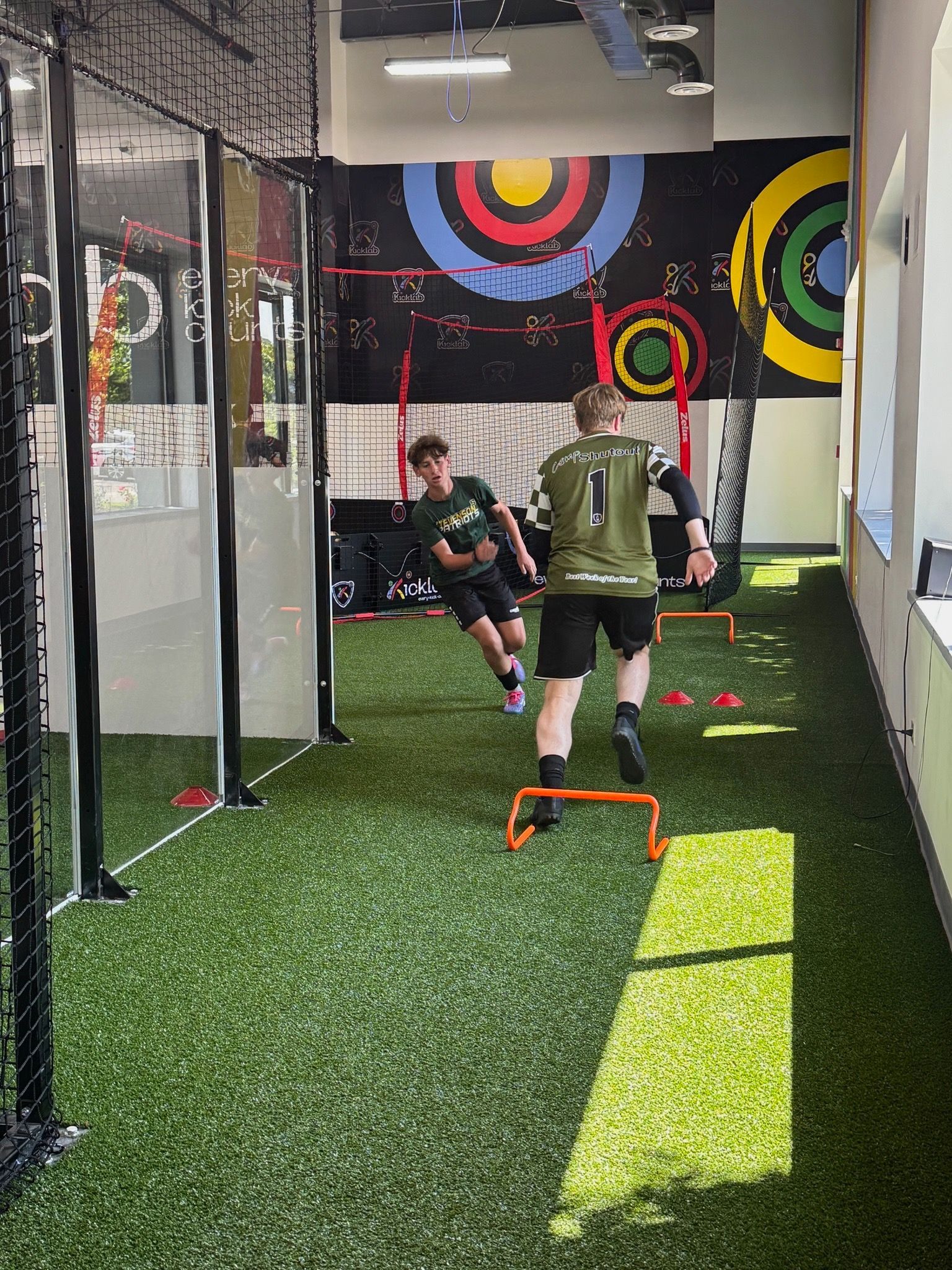 Two men training on turf, one jumping hurdles, the other running, inside a gym with colorful target art on the wall.