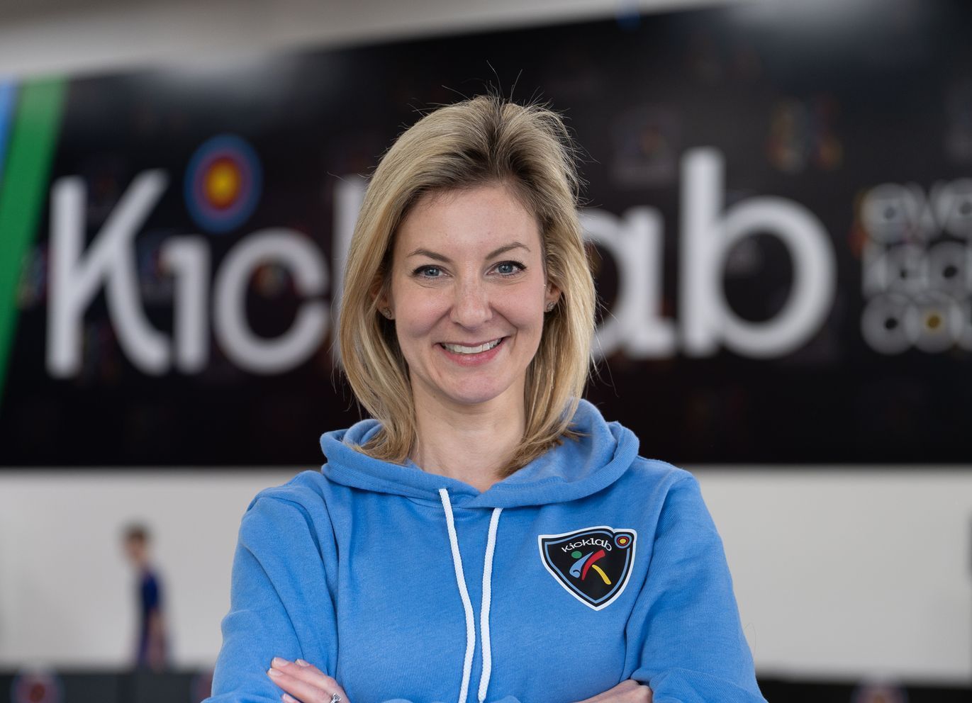 A woman in a blue hoodie is standing with her arms crossed in front of a sign that says kickab