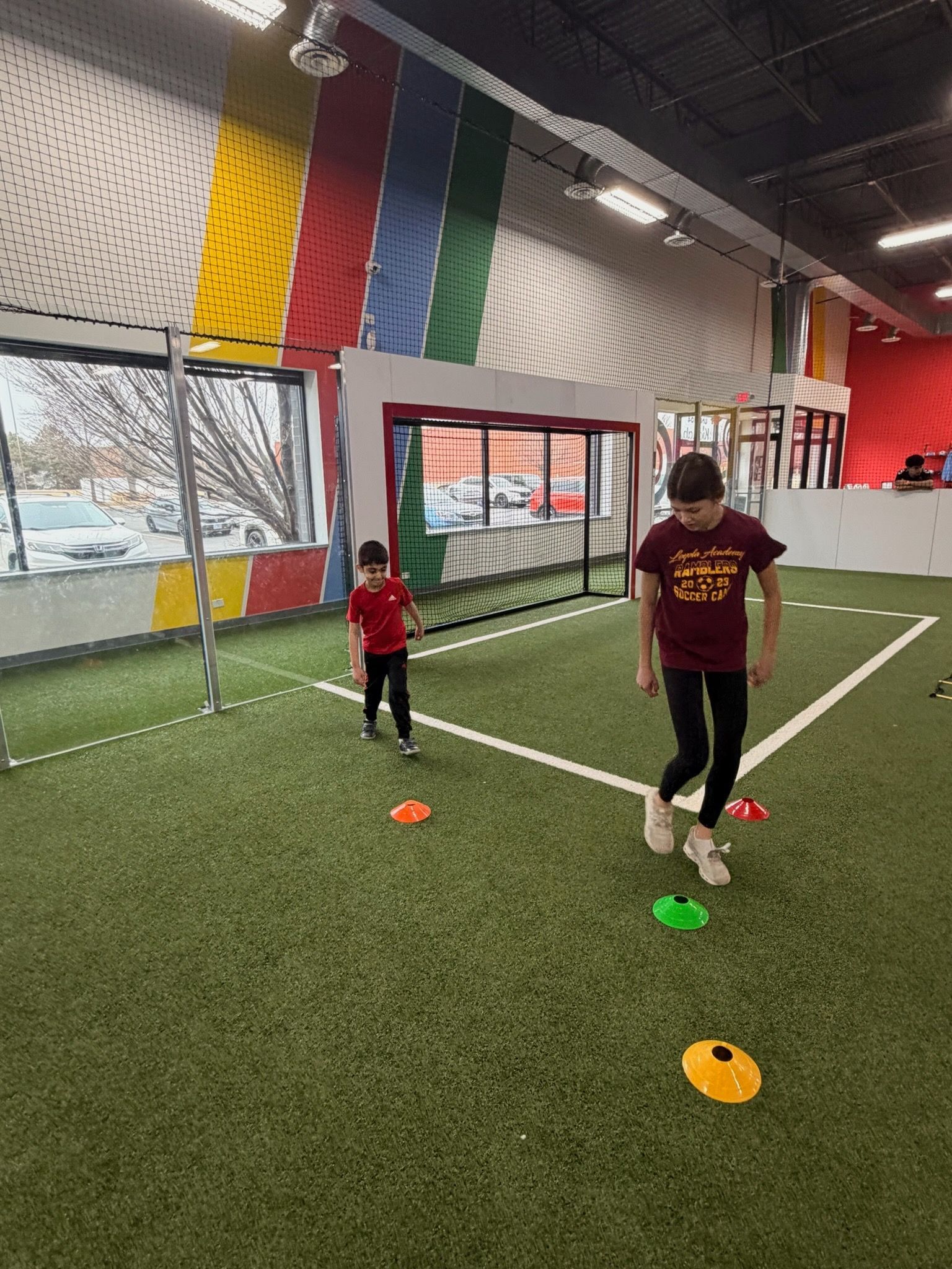 Two children in athletic clothes perform agility exercises on an indoor turf field with scattered colored cones.
