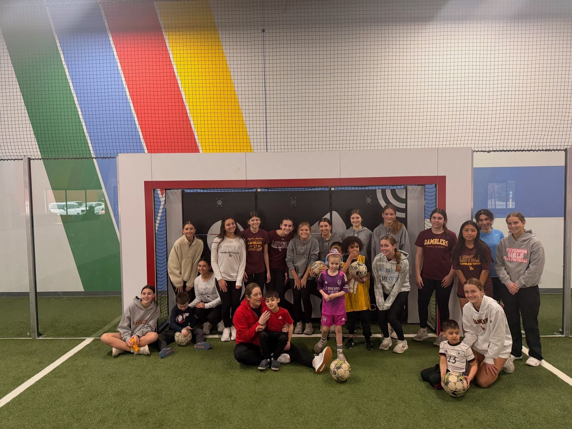A group poses for a photo in front of a soccer goal indoors, set against a wall with colorful, diagonal stripes.