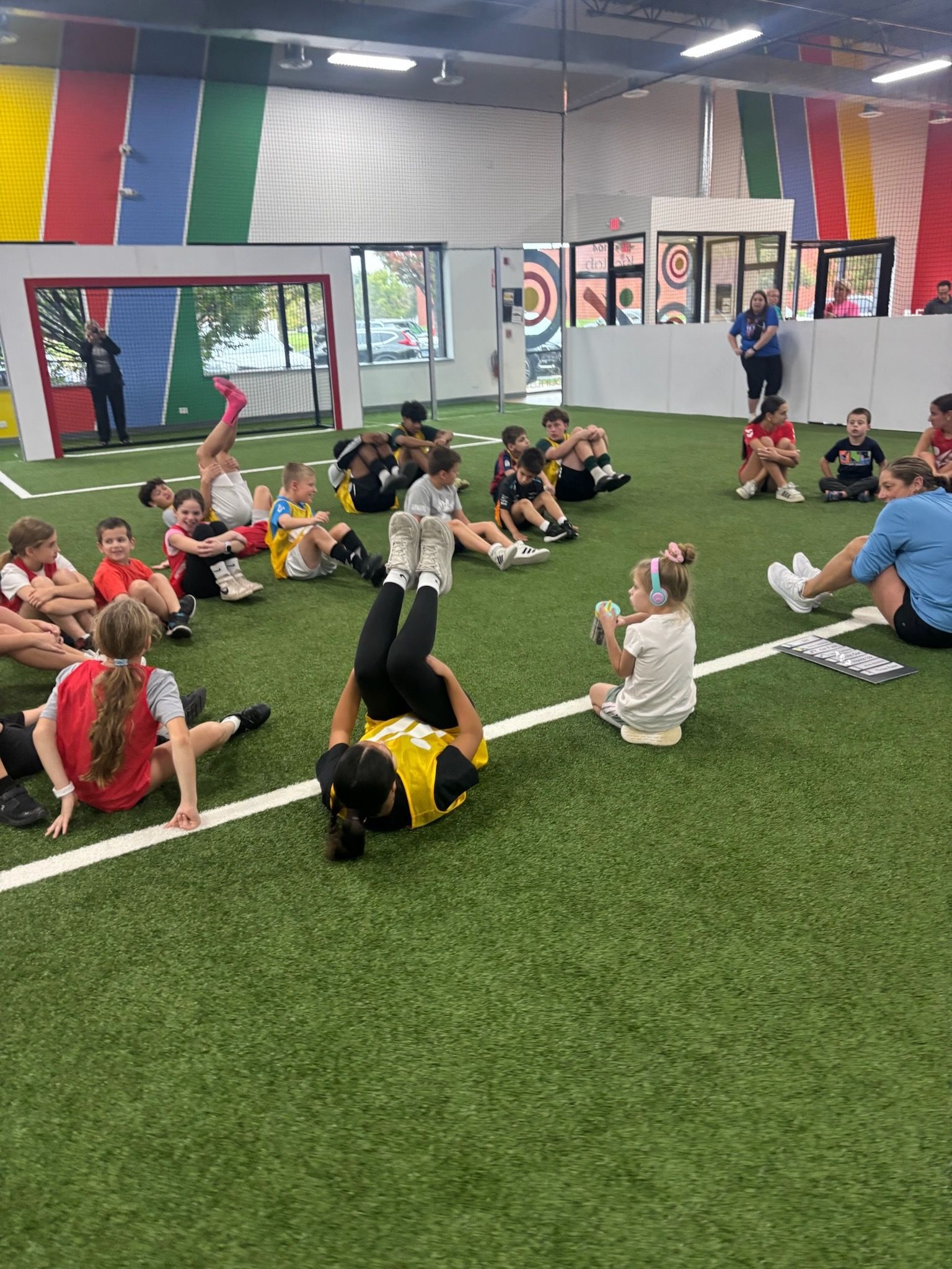 A group of children sit on an indoor turf field doing sit-ups during a sports activity.