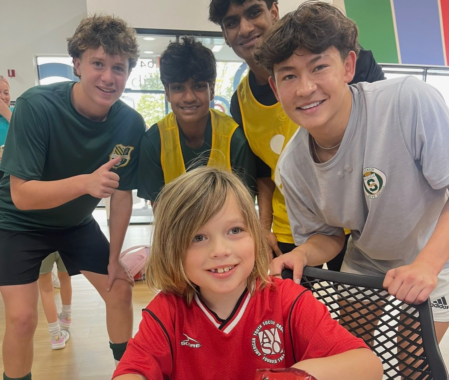 A group of five people in athletic gear smiling together indoors, with one person in a red jersey sitting in the front.