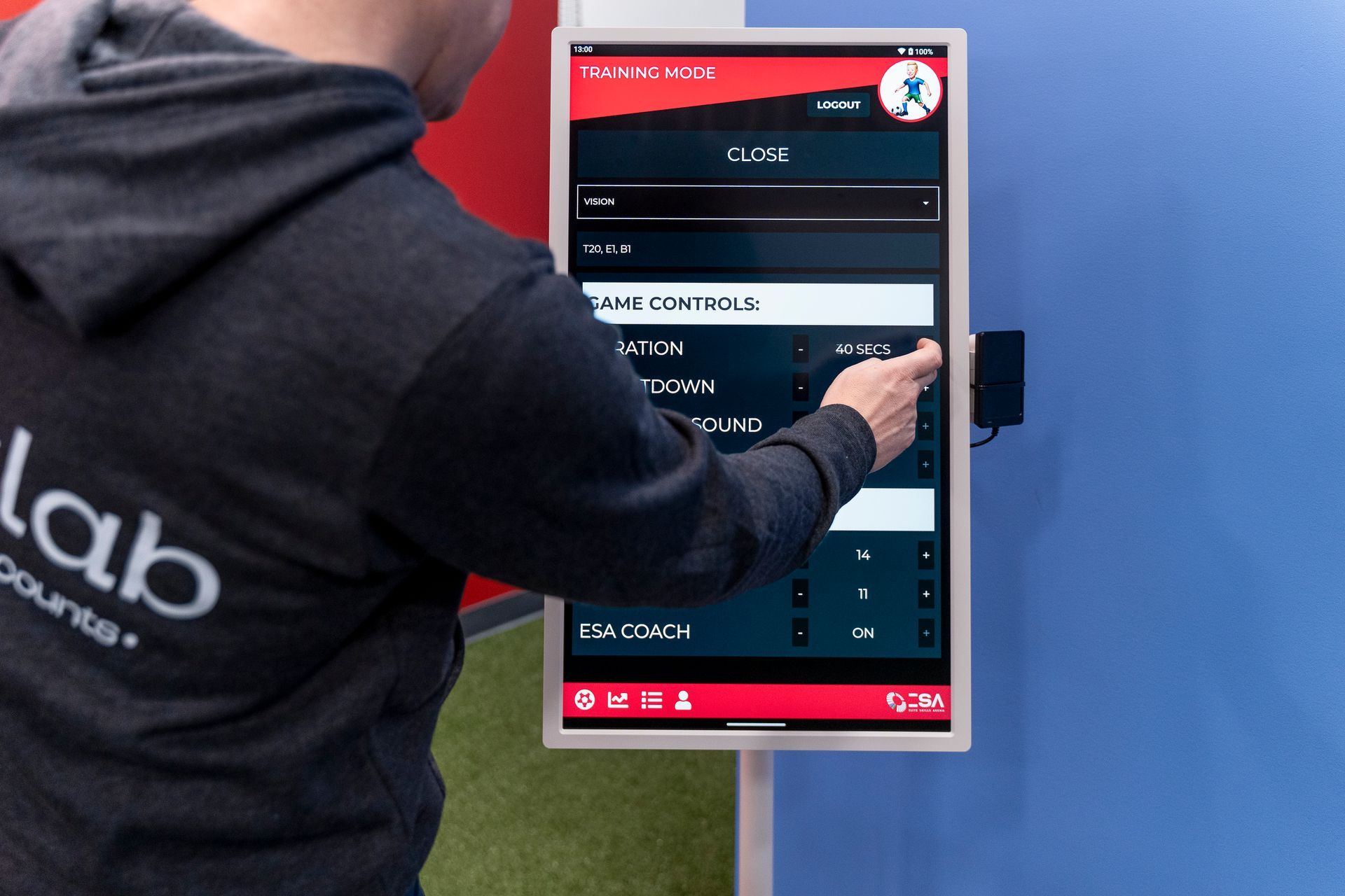 Person in gray hoodie interacting with a touch screen mounted on a blue wall. The screen displays menu options.